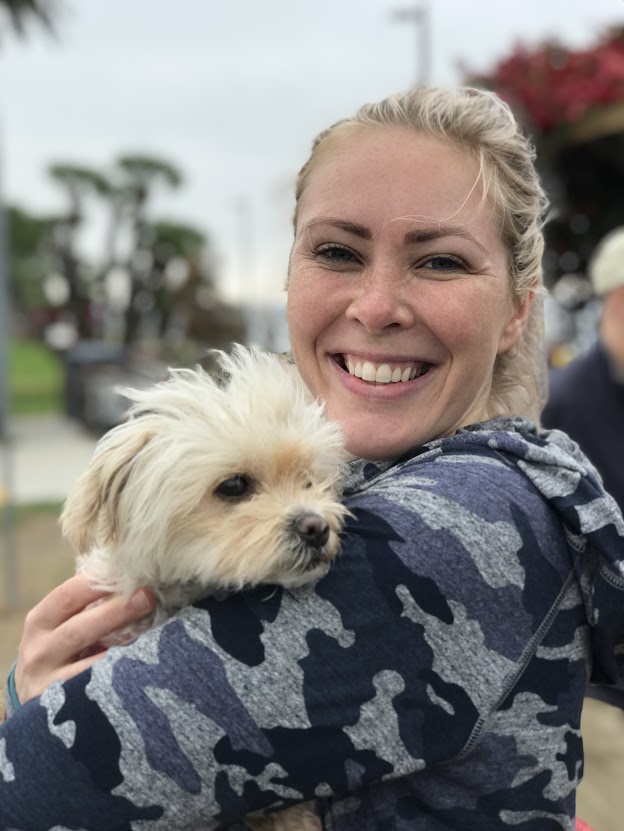 A smiling woman with long hair in a camo hoodie, holding a small fluffy dog in her arms.