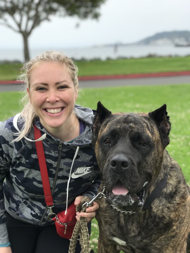 A woman smiling happily next to a large, brindle-coated dog on a grassy field, with a blurred waterfront view in the background.