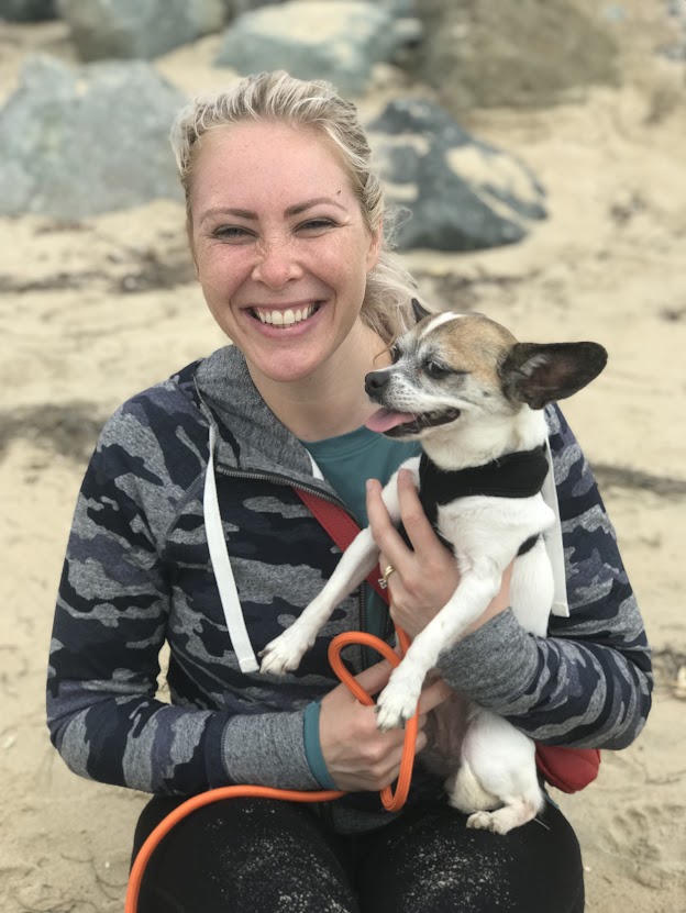 A smiling woman holds a small dog, both sitting on a sandy beach with rocks in the background.