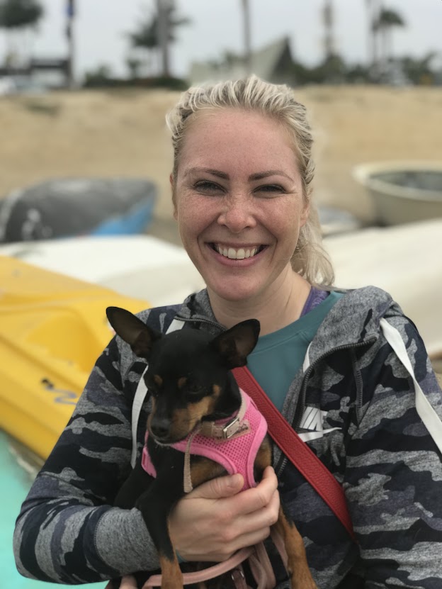 A smiling woman with blonde hair holds a small dog in a pink harness, standing in front of colorful kayaks and a sandy beach.