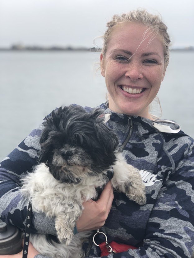 Smiling woman holding a small black and white dog, standing outdoors near water.